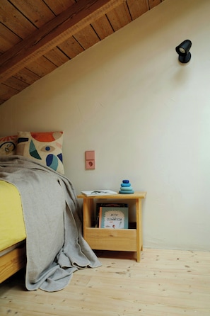 A bedroom with a wooden ceiling, a bed with a grey blanket, a bedside table with a book, and a wall-mounted light.
