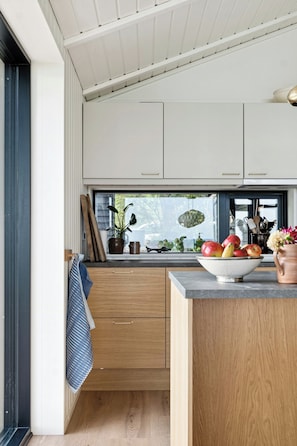 A modern kitchen with wooden cabinets, a large window, and a bowl of fruit on the counter.