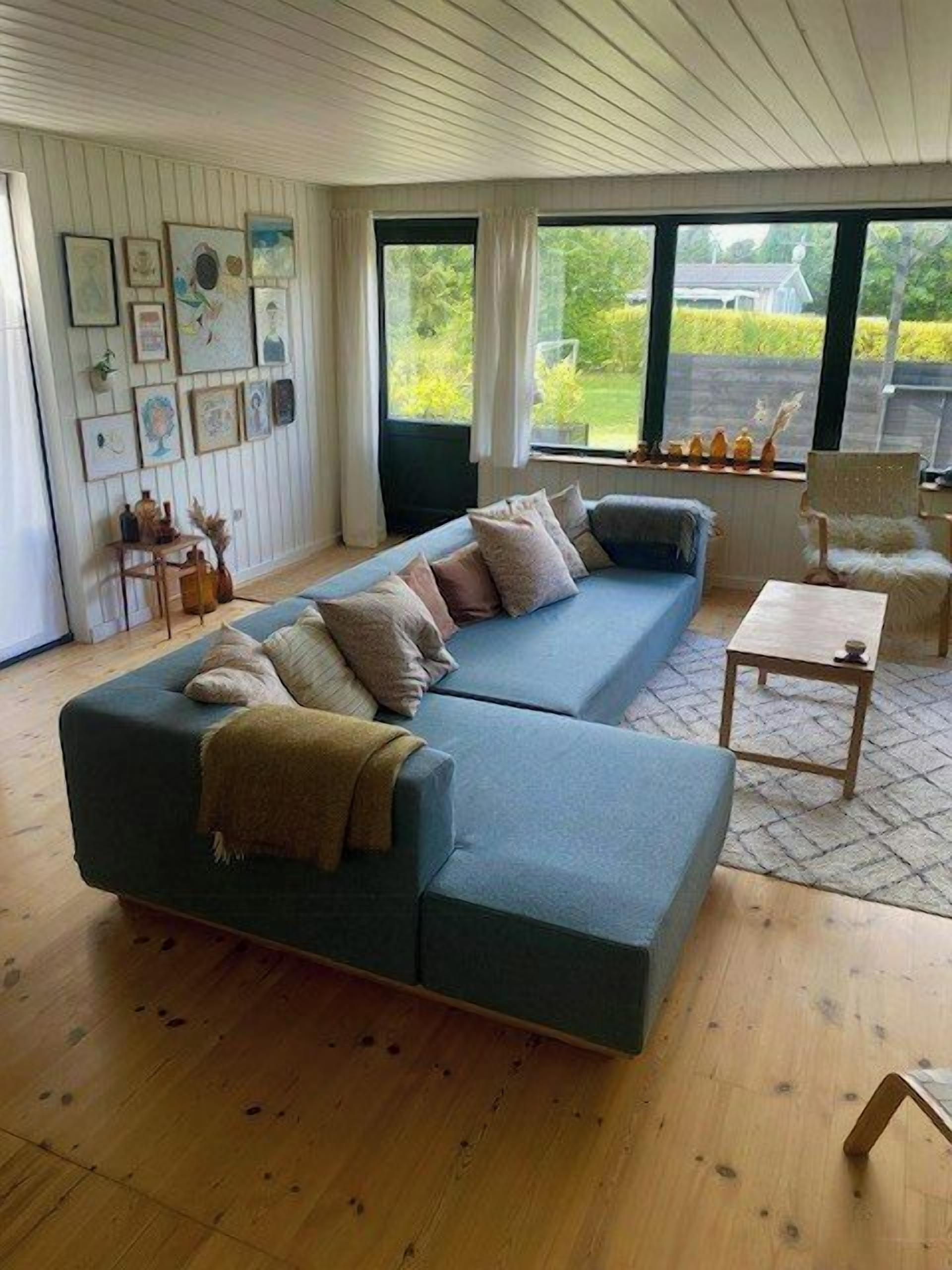 A modern living room with a sectional sofa, a coffee table, and a chair. There are framed pictures on the wall, and a window with a view of greenery outside.