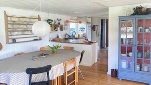 Uma sala de jantar com uma mesa redonda, cadeiras, um armário com portas de vidro e uma cozinha ao fundo.