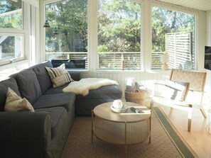 Ein gemütliches Wohnzimmer mit einem grauen Sofa, einem runden Couchtisch und einem Holzstuhl. Große Fenster bieten Blick auf Bäume und einen Balkon.
