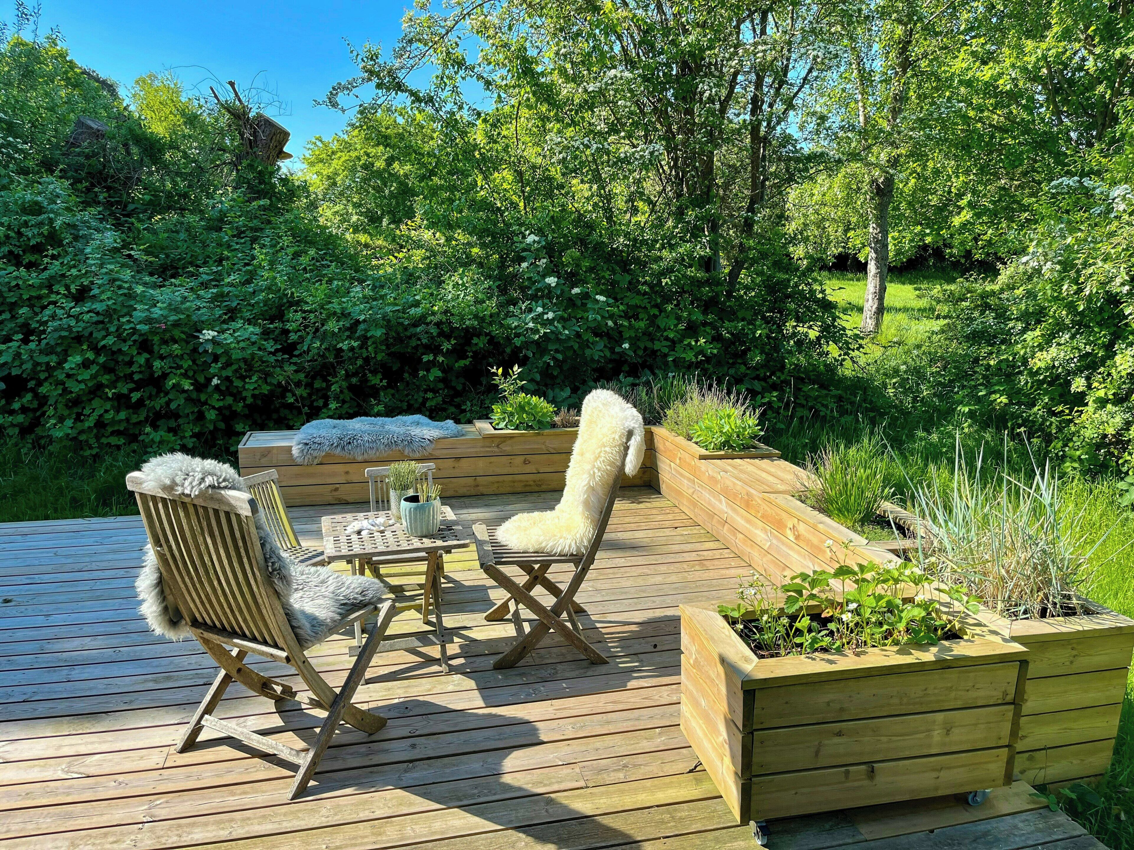 Een houten terras met tuinmeubilair, een plantenbak en een weelderige tuin op de achtergrond.