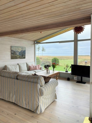 A modern living room with a large window, a sofa, a coffee table, and a painting on the wall. - Holiday home with pool and fjord views on Lundø (Højslev)