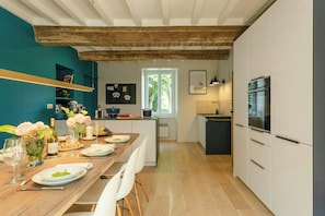 A modern kitchen with wooden beams, a dining area with a wooden table set for a meal, and a view of the outdoors through a window.