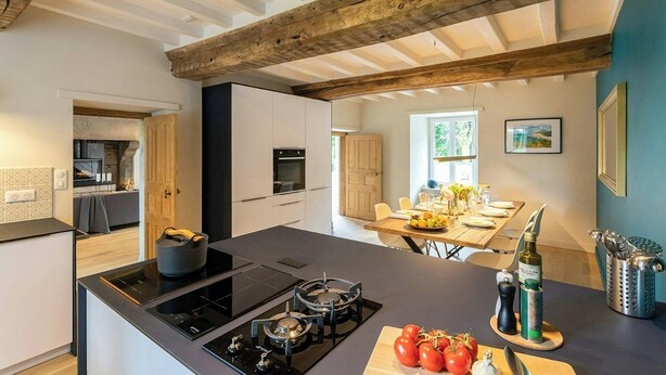 A modern kitchen with a central island, built-in appliances, and a dining area with a table set for two. - Renovated stone house in Normandy (Percy en Normandie)