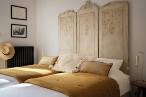 Ein Doppelbett mit Kopfteil, zwei gerahmte Bilder an der Wand, ein Hut auf der Heizung und eine Nachttischlampe.