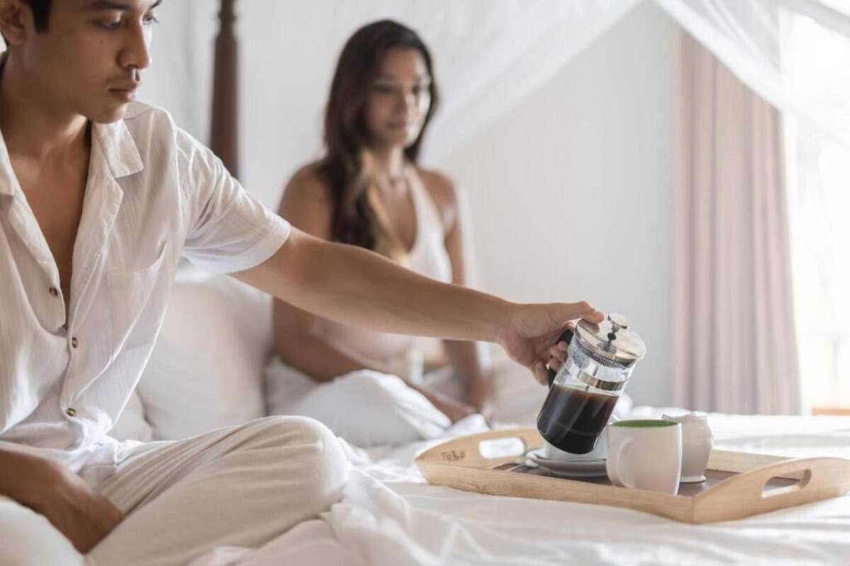 Ein Schlafzimmer mit einem Bett, einem Tablett mit Tasse und Kaffeekanne, und einer Person, die Kaffee einschenkt.