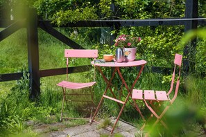 Une table et des chaises en métal de couleur rose sont disposées à l’extérieur, accompagnées d’une plante en pot et d’une bouilloire.