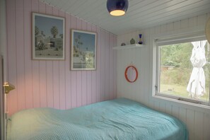 Une chambre avec un lit simple, deux tableaux encadrés au mur, une fenêtre donnant sur un paysage verdoyant et une porte blanche.