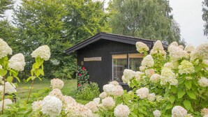 Een klein houten vakantiehuisje met een bord waarop 'Xlarmenborg 8' staat, omringd door weelderige begroeiing en witte hortensia's.