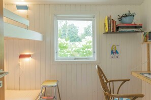 Et værelse med et vindue, en stol, et lille skamle, en hylde med bøger og en skål, samt en lampe på væggen.