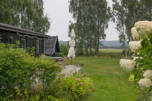 Una construcción de madera con terraza, rodeada de vegetación exuberante y un campo cubierto de césped.