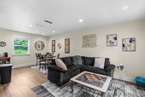 A living room with a sectional sofa, a coffee table, a dining area with chairs, and artwork on the walls.