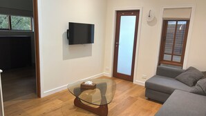 Un salón con una mesa de centro de vidrio, un sofá gris, un televisor de pantalla plana montado en la pared y una puerta de madera.
