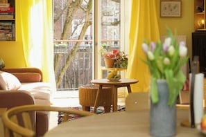 Um quarto com sofá, mesa com um vaso de flores, cadeira de vime, janela com cortinas e uma estante de livros.