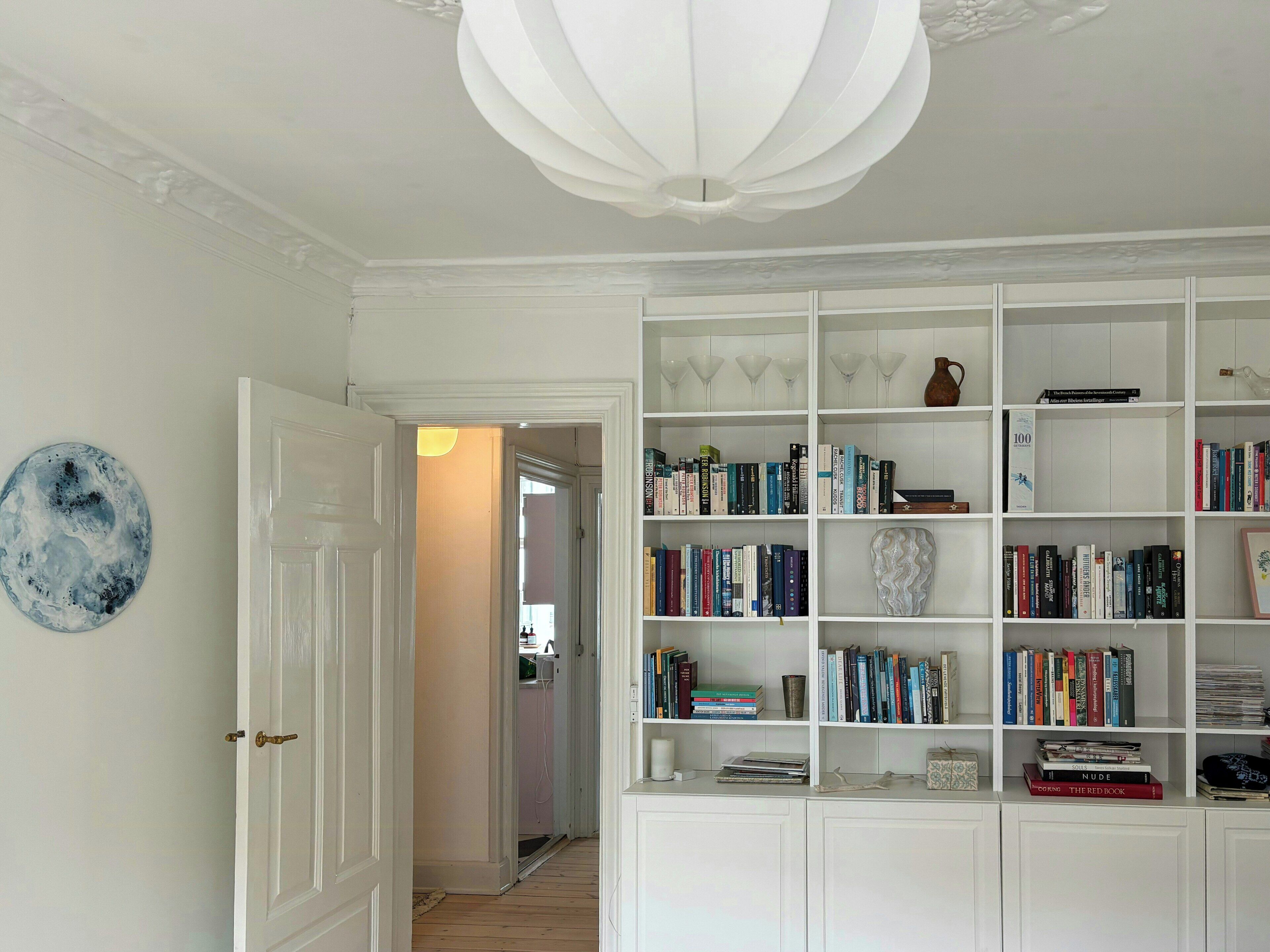 En gang med en hvid dør, et hvidt loft med en dekorativ lampe og et hvidt reolsystem fyldt med bøger og pyngeting.