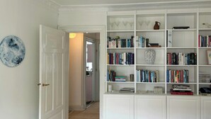 Un couloir avec une porte blanche, un plafond blanc orné d’un luminaire décoratif et une bibliothèque blanche remplie de livres et d’objets décoratifs.