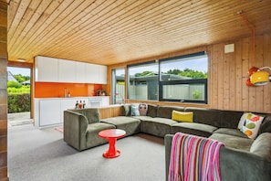 Un intérieur moderne avec un plafond en bois, une cuisine dotée de meubles blancs, un canapé d’angle gris et une table rouge.