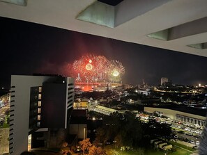 Ein beleuchteter Stadtblick bei Nacht mit Feuerwerkskörpern in der Ferne, modernen Gebäuden und einem Parkplatz.