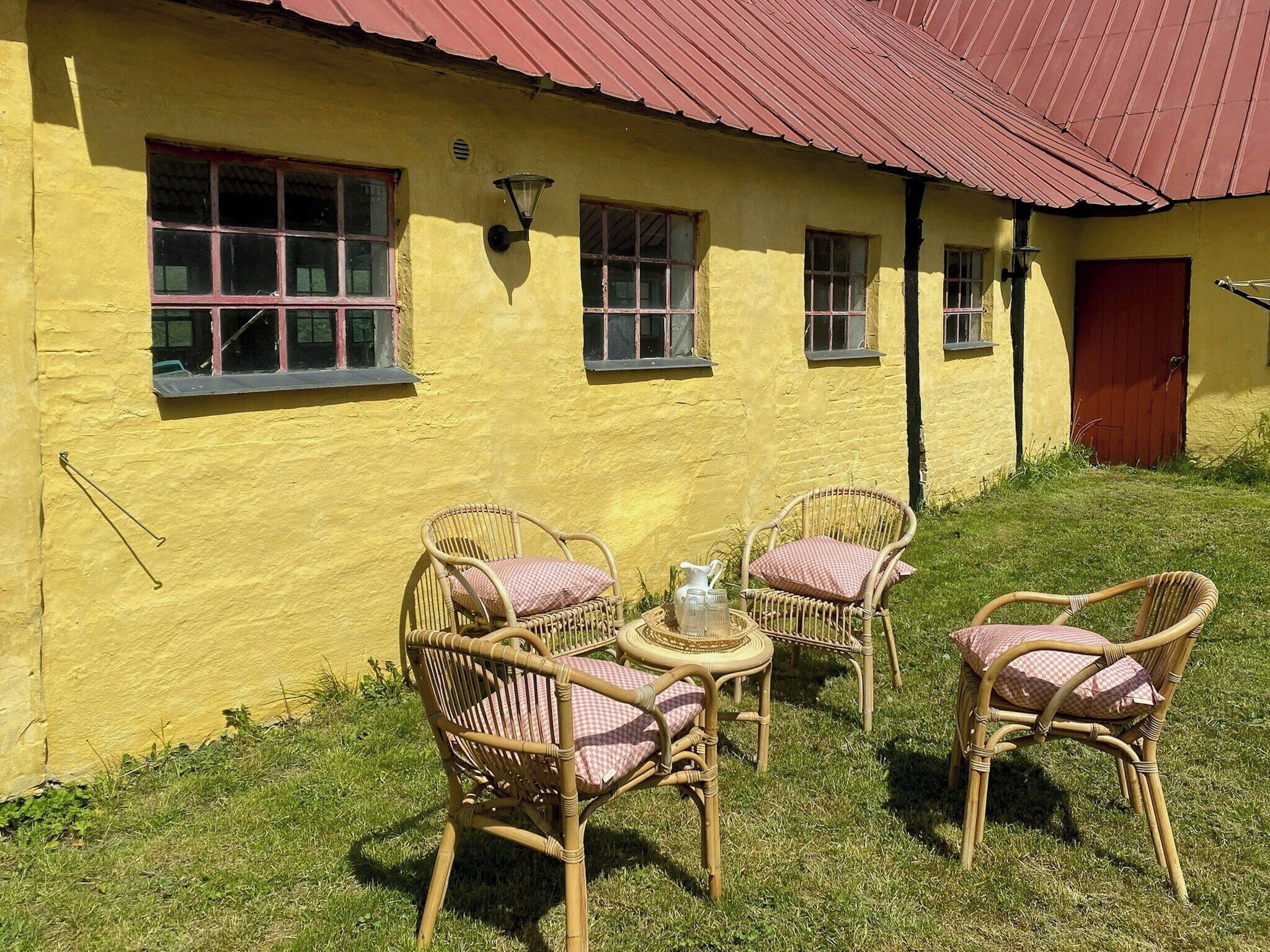 A yellow building with a red roof, wicker chairs with checkered cushions, and a small round table with a pitcher and jug.