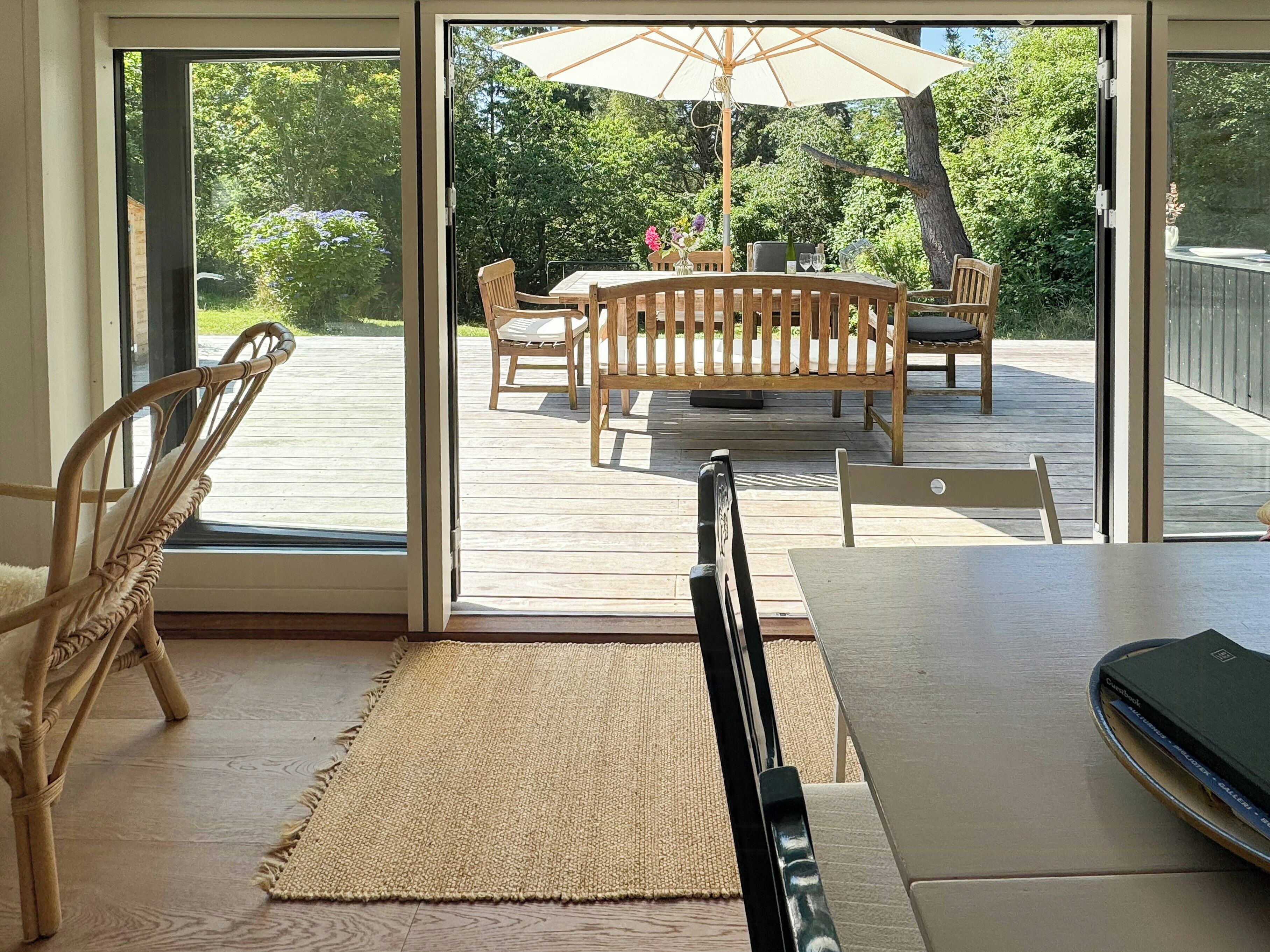 A room with a sliding glass door leading to an outdoor deck with a table and chairs, a woven rug, and a wicker chair.