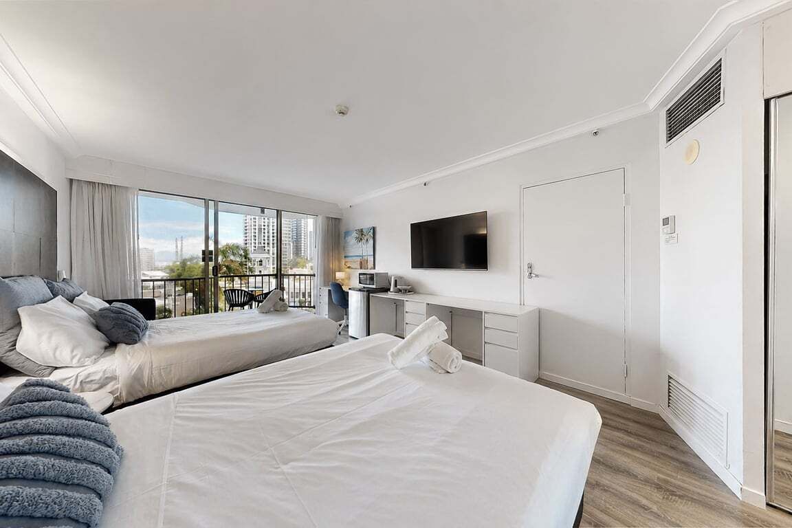 A hotel room with two beds, a TV, a desk, and a balcony with a city view.