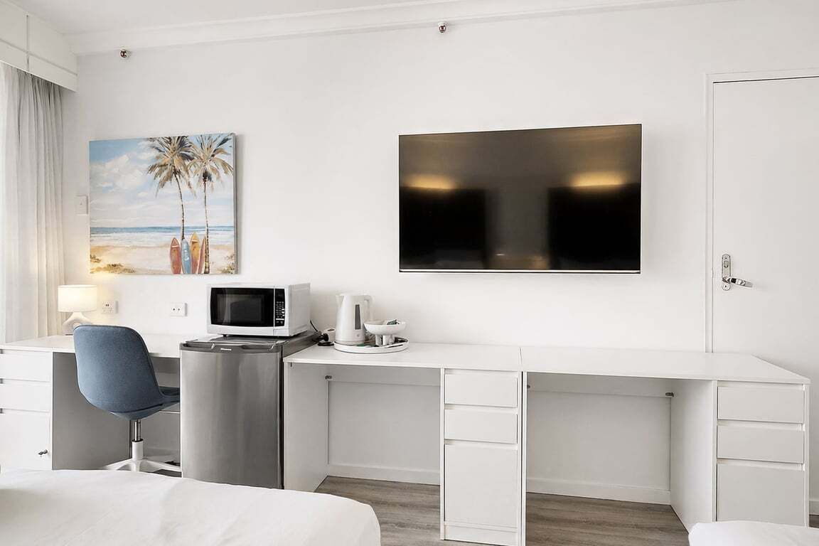 A hotel room with a flat-screen TV, a desk with a computer, a chair, a bed, and a painting of a beach scene.