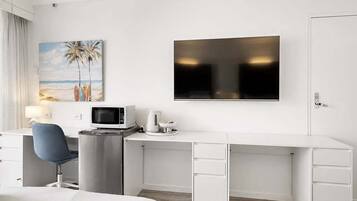A hotel room with a flat-screen TV, a desk with a computer, a chair, a bed, and a painting of a beach scene.