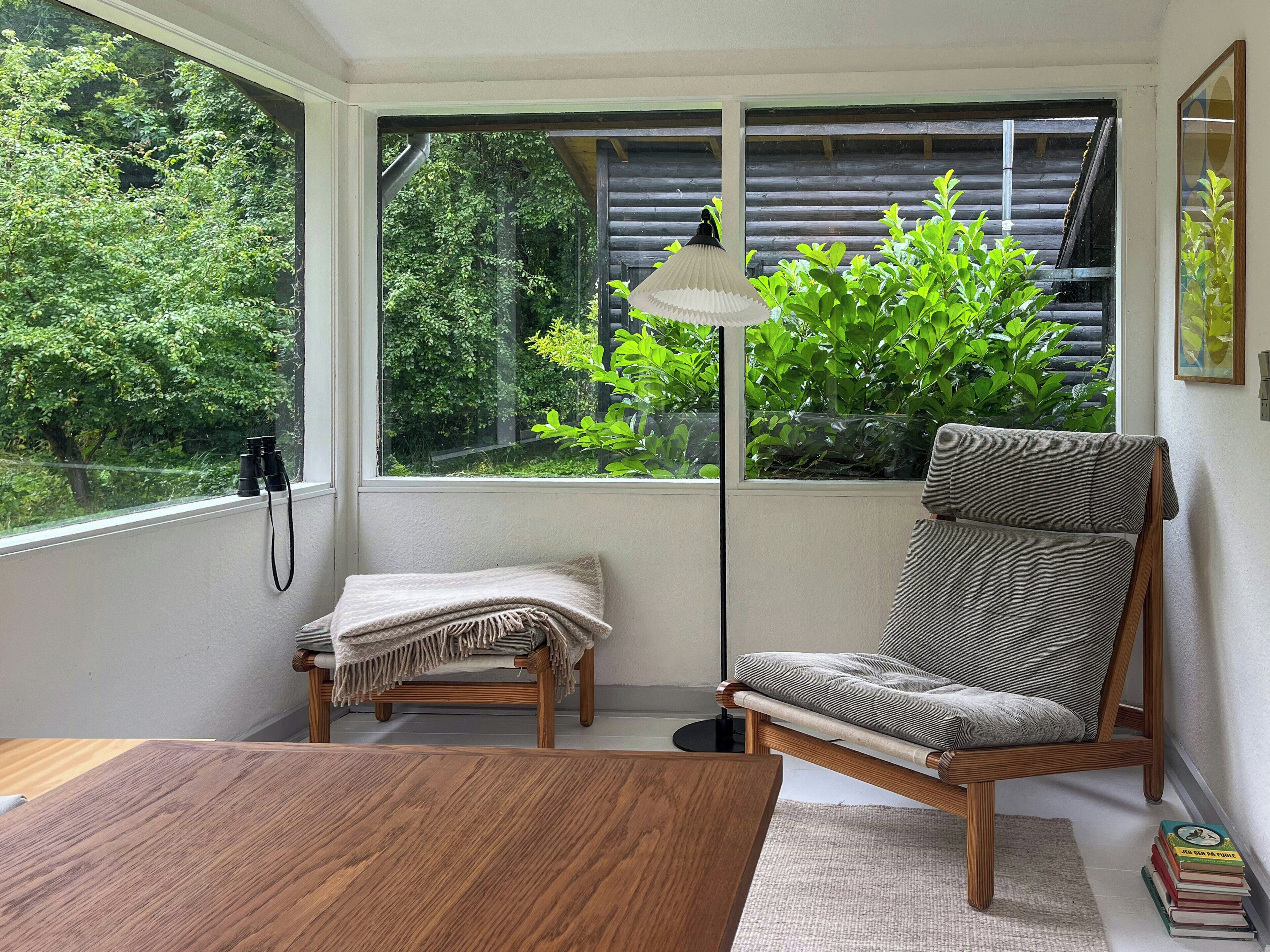 Ein Zimmer mit Holztisch, gepolstertem Sessel, Stehlampe und Blick auf Grün durch ein Fenster.