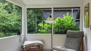Een kamer met een houten tafel, een fauteuil, een vloerlamp en uitzicht op groen door een raam.