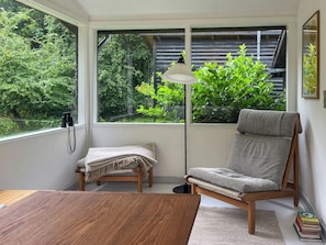 A room with a wooden table, a cushioned chair, a floor lamp, and a view of greenery through a window. - Nature-rich 60s holiday home by Isefjord (Jægerspris)