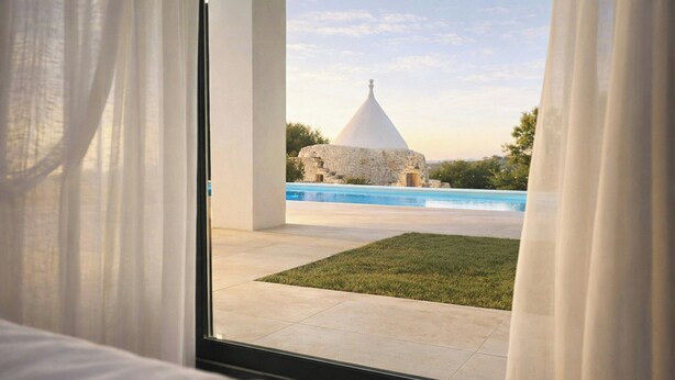Vista desde una ventana con cortinas, un edificio de piedra y una piscina.