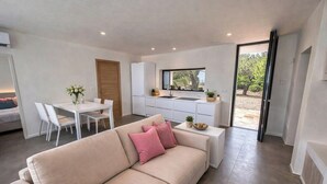 Una sala de estar moderna con un sofá color beige, una mesa de comedor con sillas, una cocina con refrigerador y un cerramiento de vidrio corredizo que da a un espacio exterior.