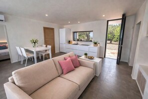 Ein modernes Wohnzimmer mit einem beigefarbenen Sofa, einem Esstisch mit Stühlen, einer Küchenzeile inklusive Kühlschrank und einer Glastür zum Außenbereich.