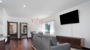 Un salon moderne avec un canapé d’angle gris, un téléviseur à écran plat, un meuble en bois et un coin repas avec des chaises.