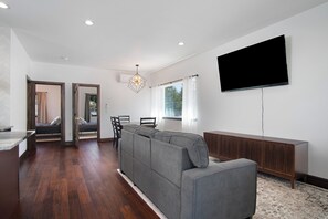 Ein modernes Wohnzimmer mit einem grauen Ecksofa, einem Flachbildfernseher, einer Holzkabine und einem Essbereich mit Stühlen.