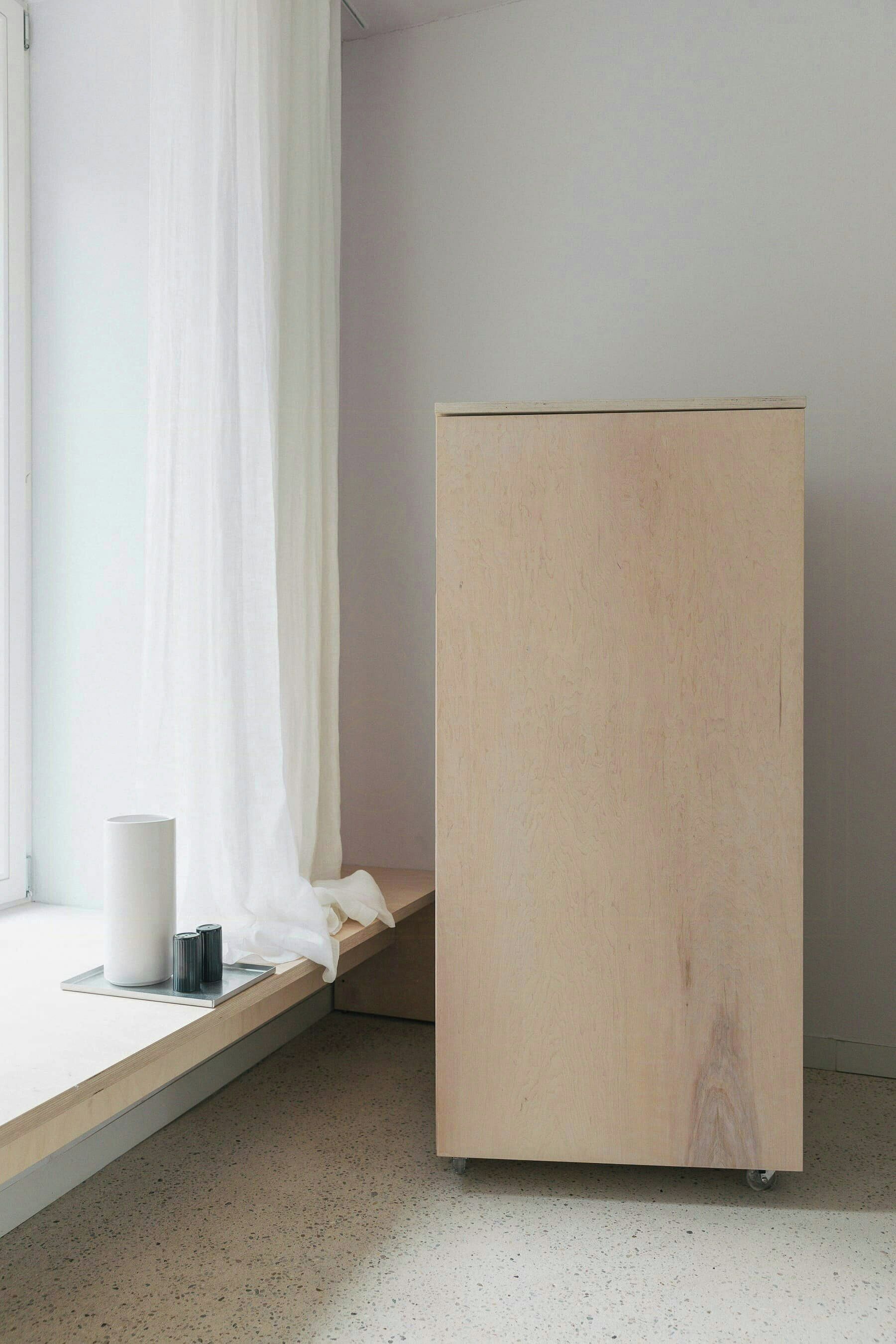 Een kamer met een houten kast, een raam met dunne gordijnen, en een tafeltje met een wit voorwerp en een zwarte houder.