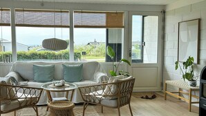 Ein Zimmer mit einem weißen Sofa, Korbstühlen, einem Glastisch und Blick ins Freie.