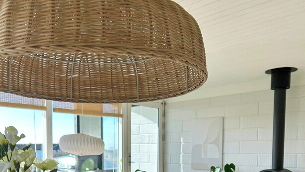 Ein modernes Wohnzimmer mit einer Korbleuchte an der Decke, einem weißen Sofa, einem Korbstuhl, einem Kamin und einem großen Fenster.