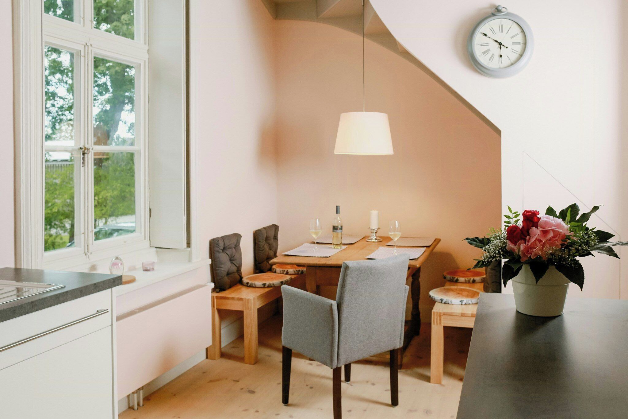 Een keuken met een eethoek, een klok aan de muur en een raam met uitzicht op groen.
