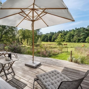 A wooden deck with outdoor furniture, a large umbrella, and a view of a green field with a fence and trees. - Design-led nature retreat, near Hornbæk beach (hornbæk)
