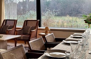 A dining area with wicker chairs, a table set for four, and a view of a forest through a large window. - Design-led nature retreat, near Hornbæk beach (hornbæk)
