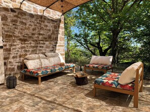 A stone-walled outdoor seating area with two wooden sofas and a chair, a woven basket, and a bottle on a wooden table. - House with swimming pool in Monts d'Ardèche (GRAVIERES)