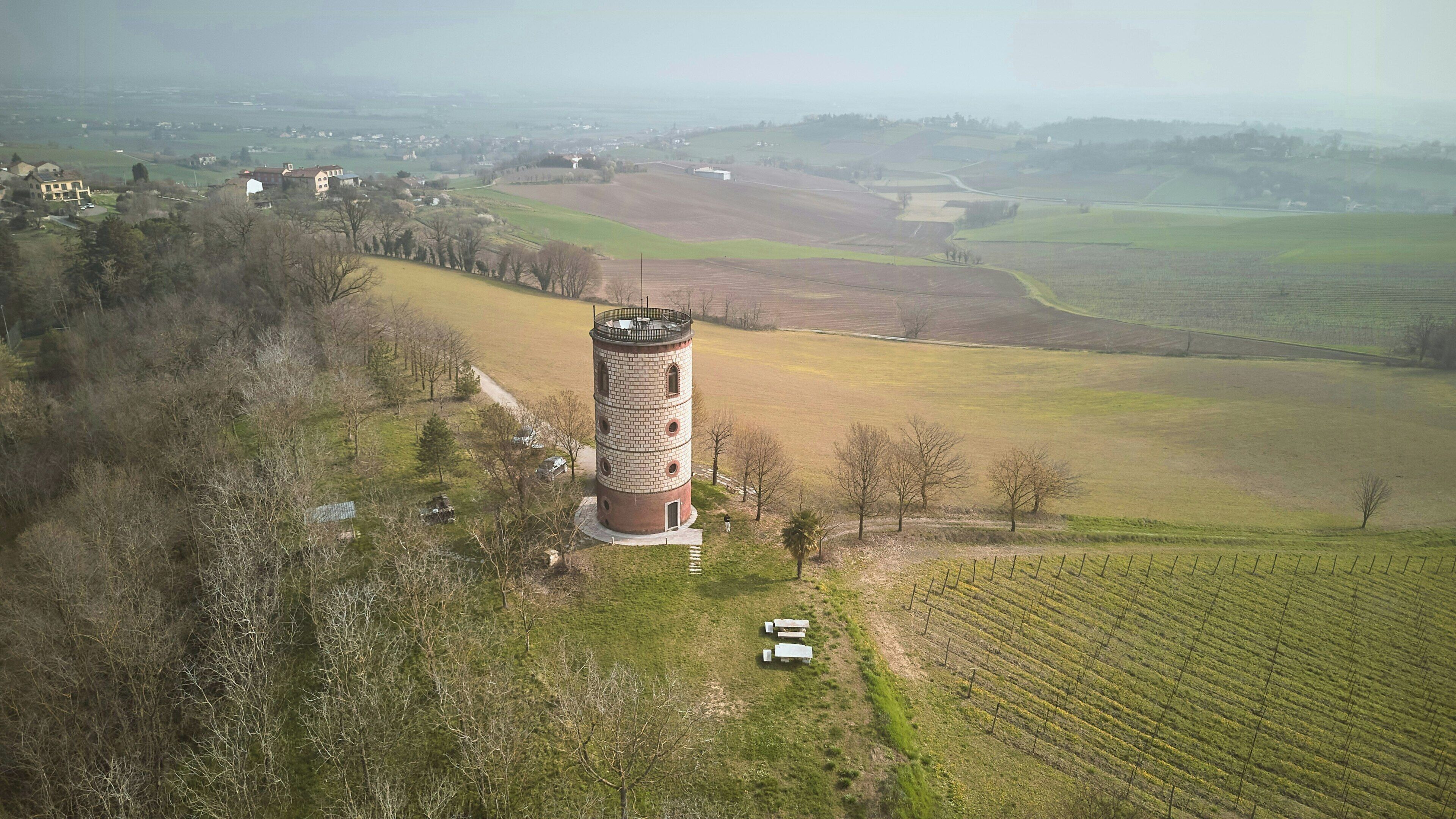 An aerial view of a tower with a brick base and a cylindrical top, surrounded by fields and sparse trees.