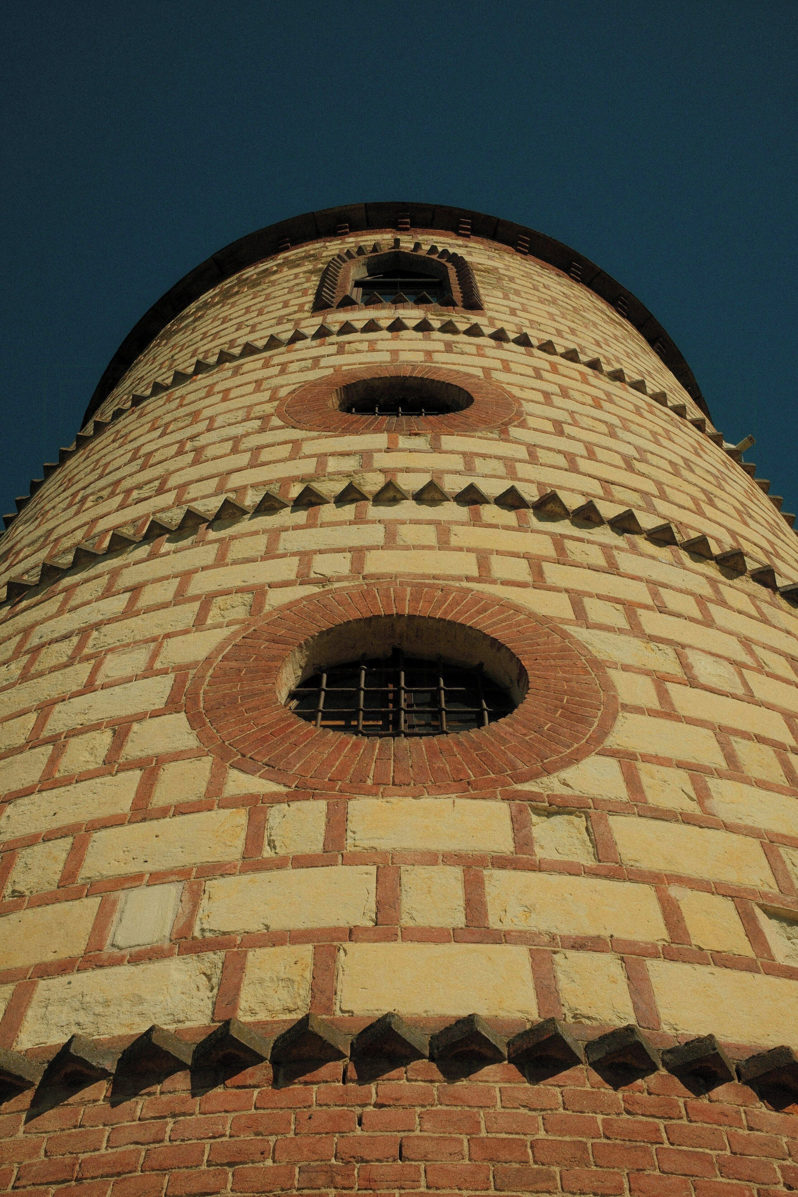 Une haute tour cylindrique en briques, dotée de fenêtres rondes et surmontée d’un toit en forme de pyramide.