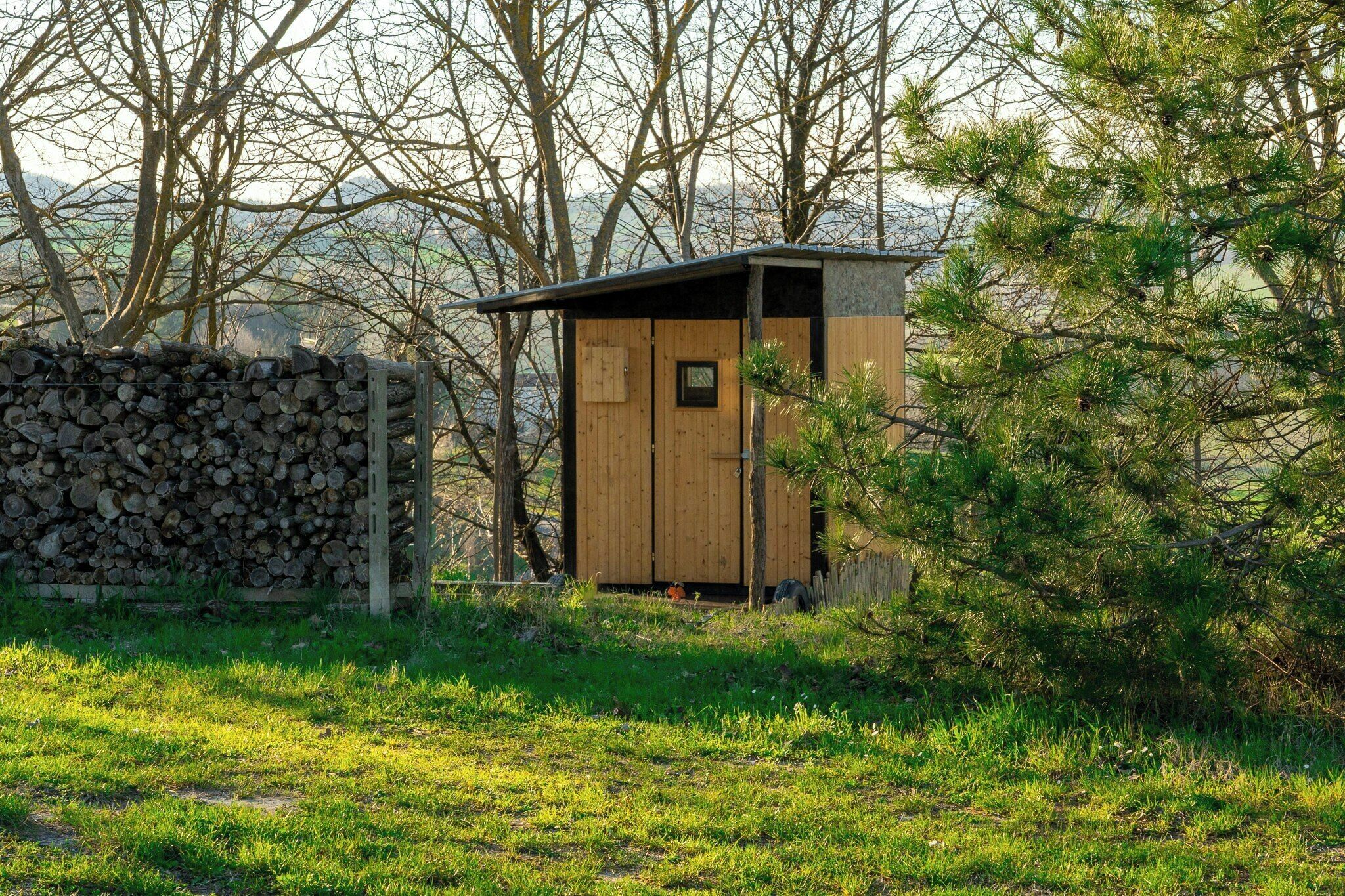 Eine kleine Holzhütte mit geneigtem Dach, gelegen in einer grünen Wiese mit Bäumen im Hintergrund.