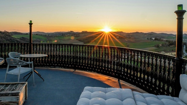 A balcony with a view of the sunset, a white cushioned seating area, and a metal railing. - Historic tower with Monferrato views (Terrugia)