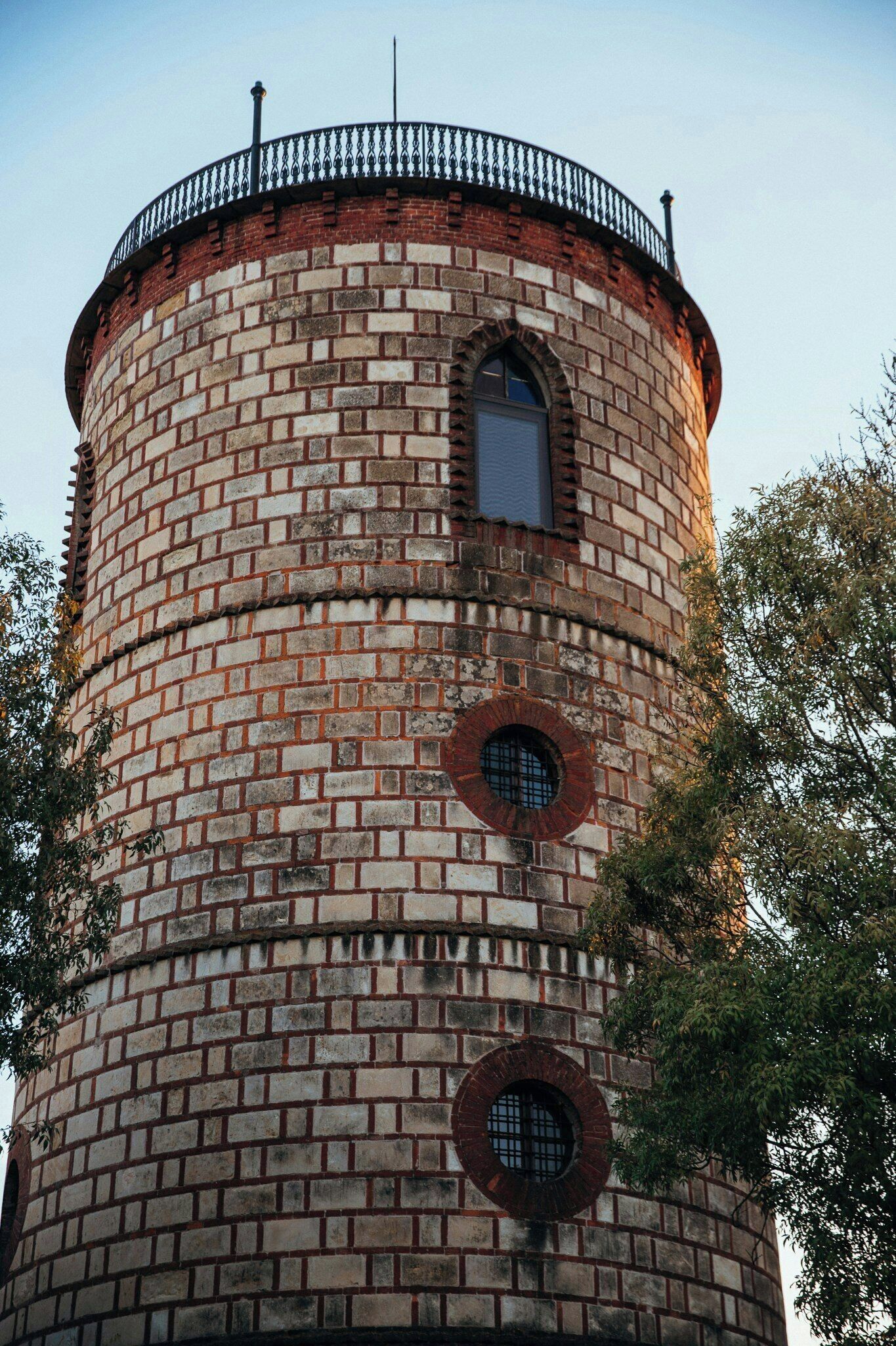 A cylindrical brick tower with a metal railing at the top, a small arched window, and two circular windows.