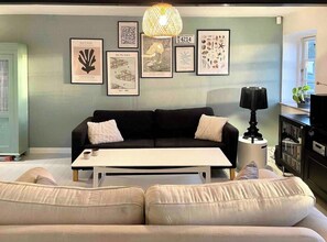 Un salon moderne avec un canapé noir, une table basse blanche et un mur décoré d’œuvres d’art encadrées.