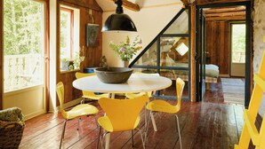 A wooden interior with a round table, yellow chairs, a large black pendant light, and a view through a glass door.